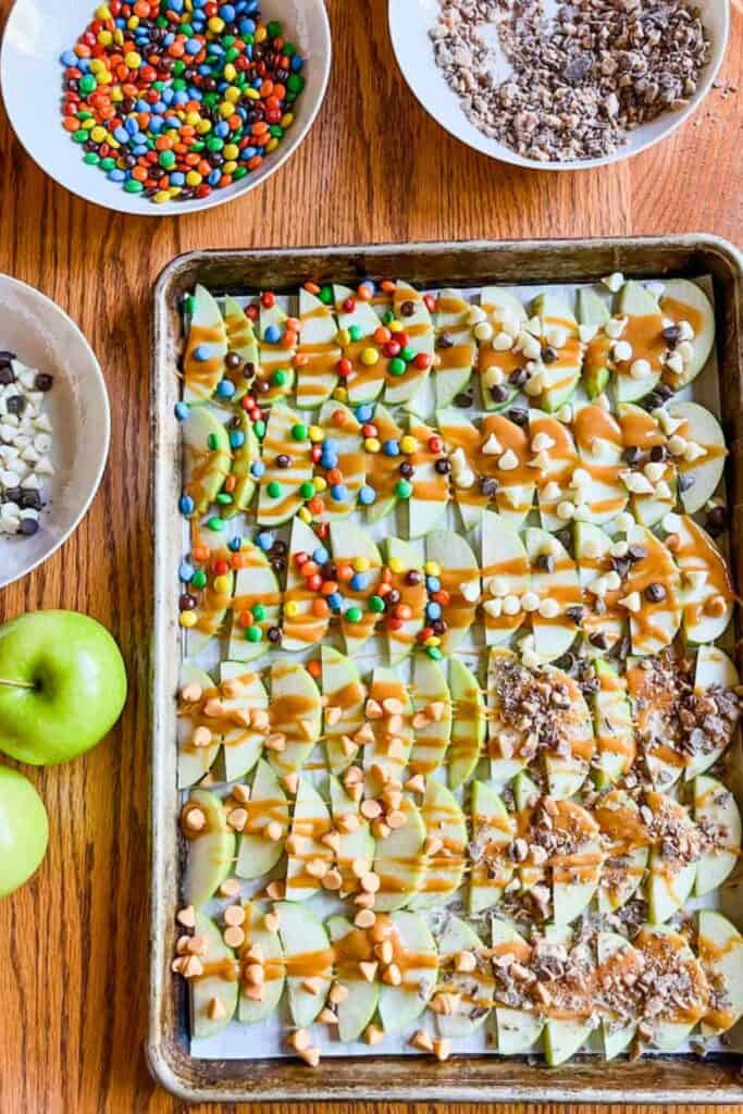 Sheet pan caramel apples made with rows of sliced green apples drizzled with caramel and topped with colorful candy pieces, chocolate chips, butterscotch chips, and crushed candy bars. Bowls of assorted toppings and whole green apples surround the tray.