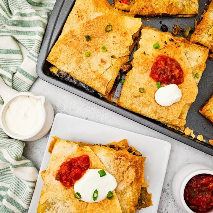 Overhead view of golden sheet pan beef quesadillas cut into squares, topped with sour cream, salsa, and green onions. One serving is plated with extra salsa and sour cream on the side, showcasing a crispy tortilla exterior and savory filling.