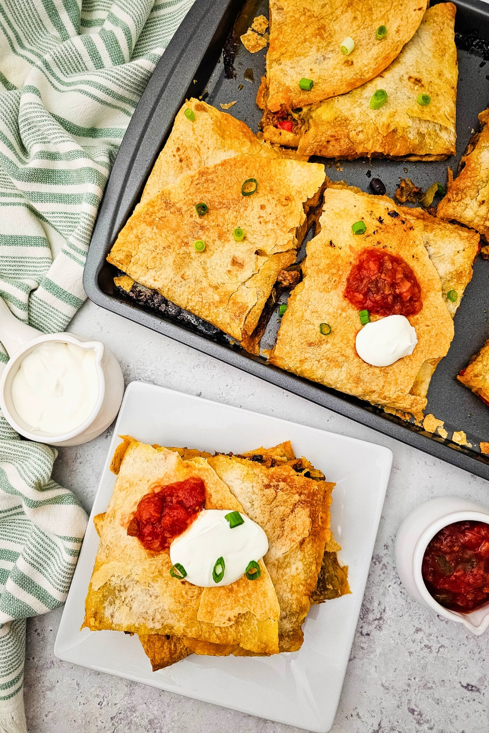 Overhead view of golden sheet pan beef quesadillas cut into squares, topped with sour cream, salsa, and green onions. One serving is plated with extra salsa and sour cream on the side, showcasing a crispy tortilla exterior and savory filling.