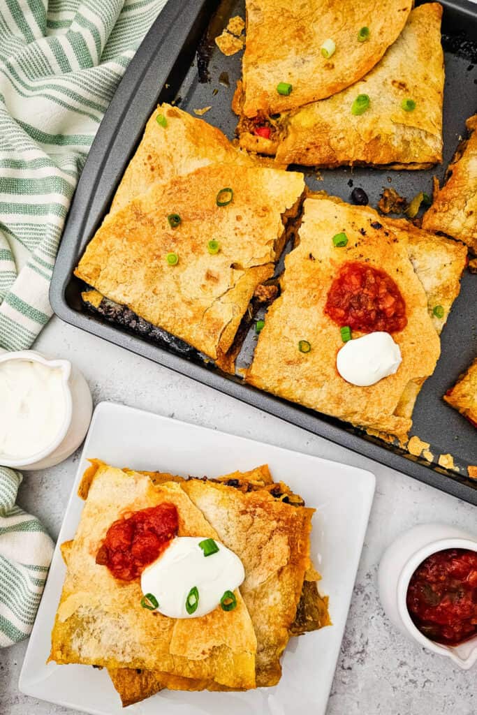 Overhead view of a baked sheet pan of quesadillas cut into crispy squares, with some topped with sour cream, salsa, and chopped green onions. Served alongside a dipping dish of sour cream for a quick dinner. 