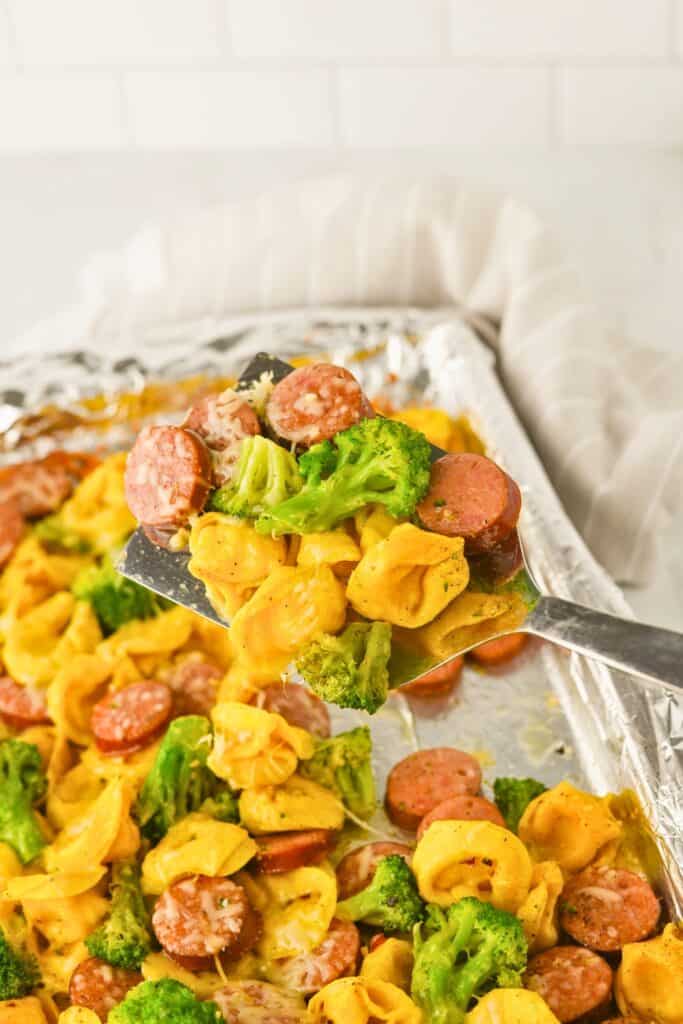 A metal spatula lifts a serving of sheet pan tortellini and sausage with melted cheese and roasted broccoli from a foil-lined baking tray. The dish is golden, cheesy, and packed with flavor, showing an easy one pan dinner straight from the oven.
