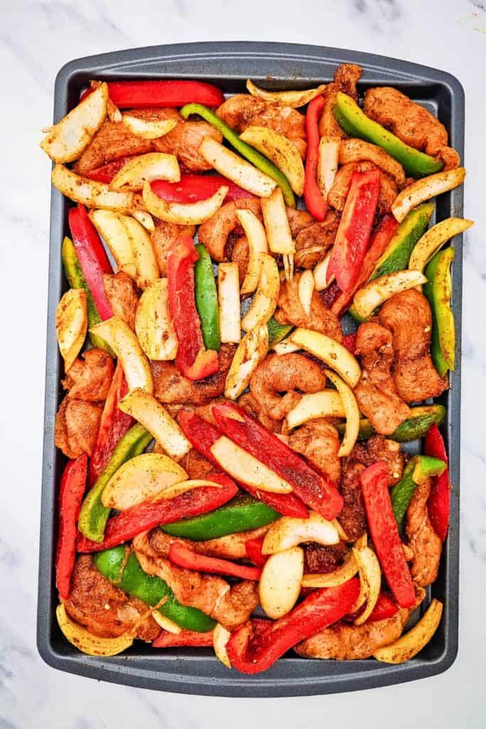 Raw chicken fajitas arranged on a sheet pan with seasoned chicken strips, sliced red and green bell peppers, and onion wedges spread in an even layer. The ingredients are ready to be baked in the oven.