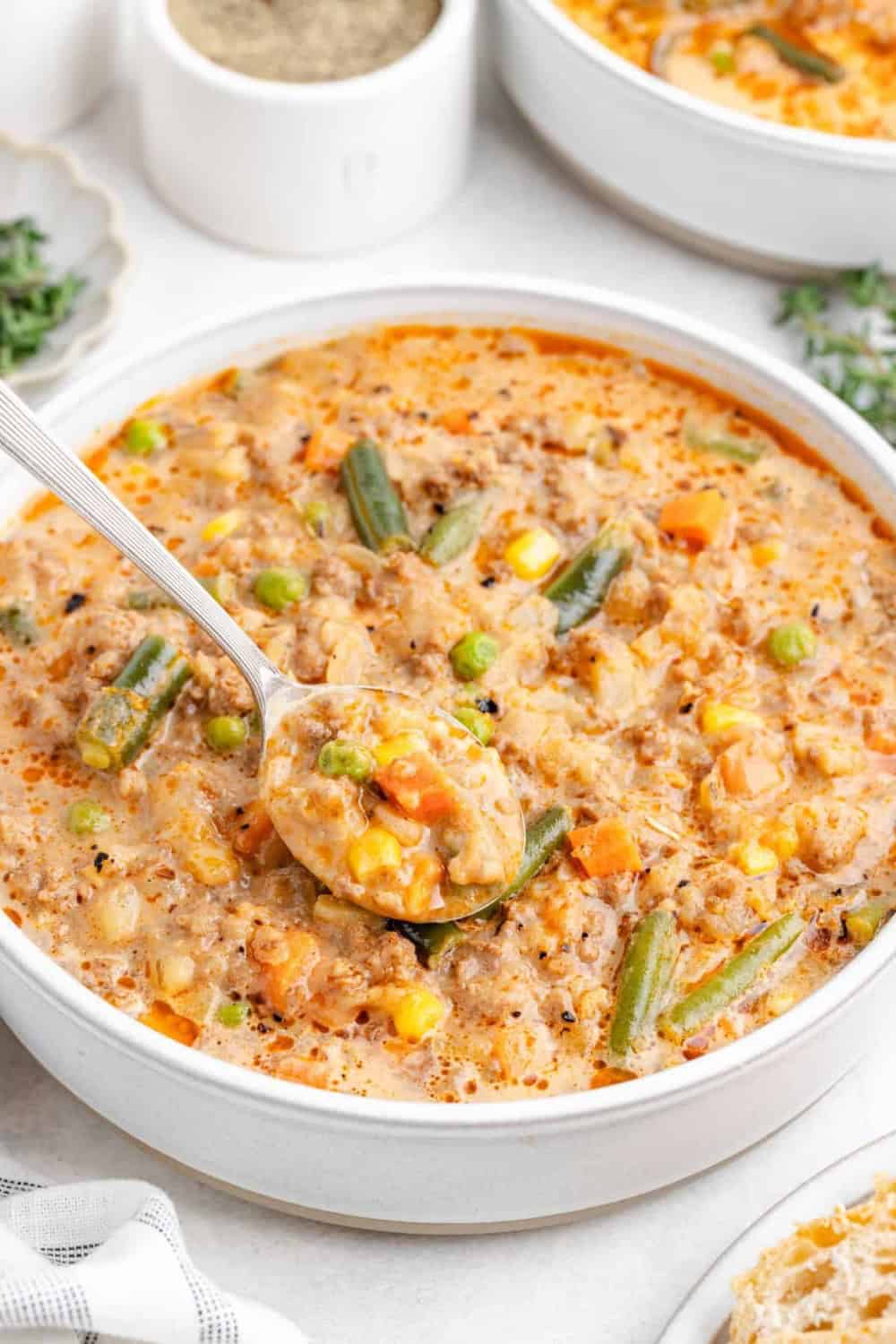 Shepherd’s pie soup with ground beef potatoes carrots peas corn and green beans is served in a creamy broth. A spoonful shows the hearty meat and vegetables in the thick soup.