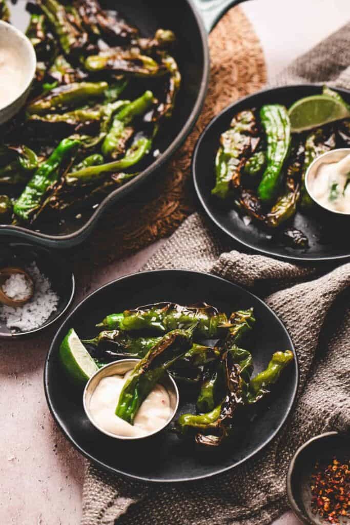 Blistered shishito peppers served on black plates with creamy dipping sauce and lime wedges, set against a rustic table with a textured brown cloth. A cast iron skillet filled with more roasted peppers sits nearby.