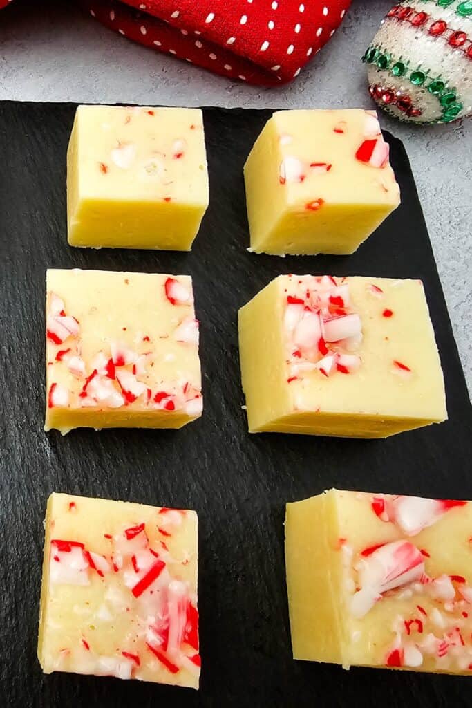 Squares of shortcut fudge topped with crushed candy canes, arranged on a black serving board with festive Christmas decorations in the background.