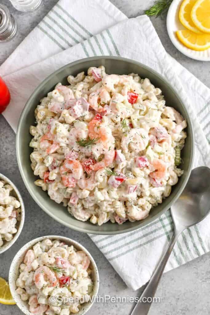 Bowl of shrimp pasta salad with elbow macaroni cooked shrimp diced red bell pepper and celery tossed in a creamy dressing and topped with fresh dill. Lemon slices and small bowls of the salad sit nearby.