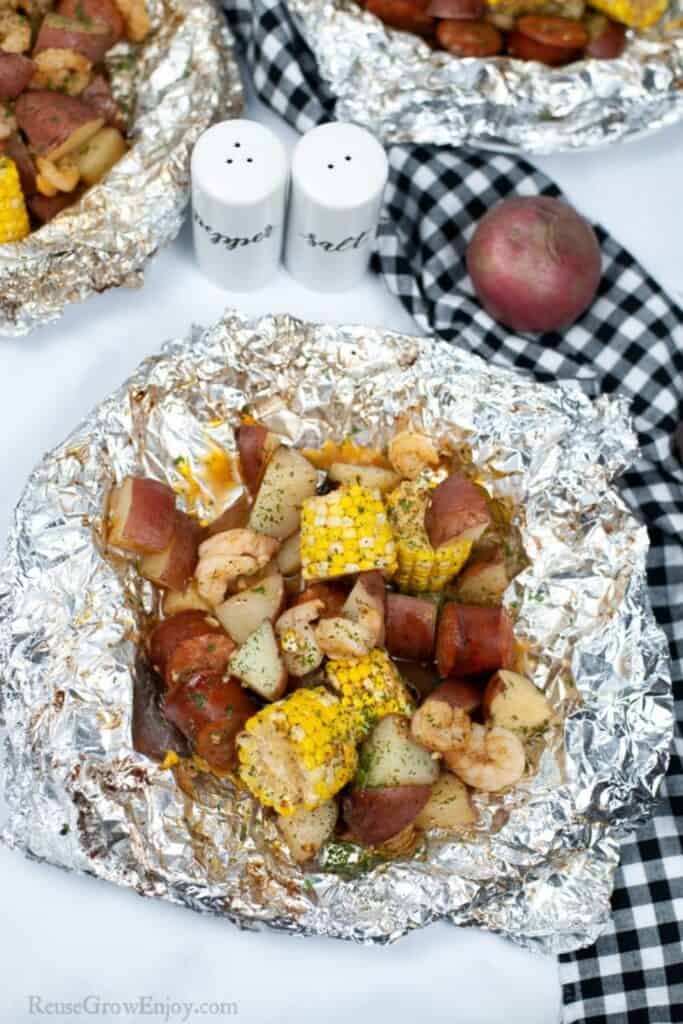 Foil pack camping dinner on a picnic table with shrimp, corn on the cob and red potato cubes. 