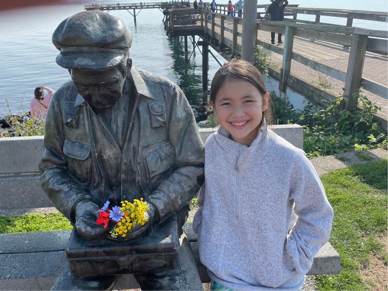 Young girl sitting beside a metal statue of a man, in front of a pier on the Si