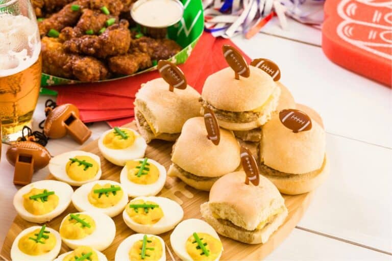 A festive spread of football Sunday appetizers including deviled eggs decorated with chive “laces,” mini cheeseburger sliders with football toothpicks, and saucy chicken wings garnished with green onions, served with a cold beer and football-themed party decor.