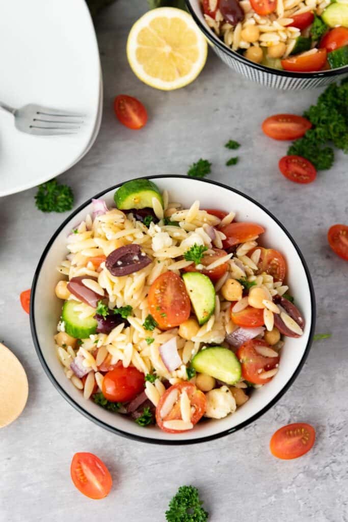 Overhead view of a medium sized round ceramic bowl of orzo salad with kalamata olives, diced red onion, feta, cherry tomato halves and chickpeas.