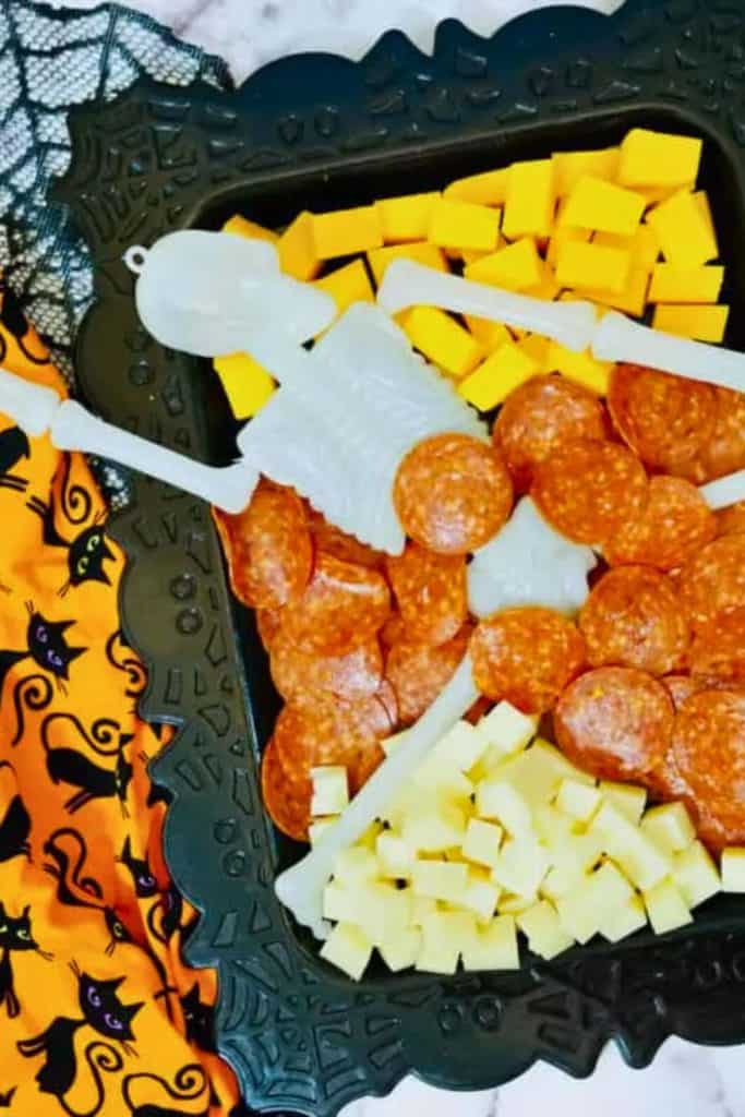Halloween charcuterie board featuring sliced pepperoni and yellow and white cheese cubes arranged around a plastic skeleton prop on a black spiderweb tray. A festive orange napkin with black cat designs adds a playful Halloween touch.
