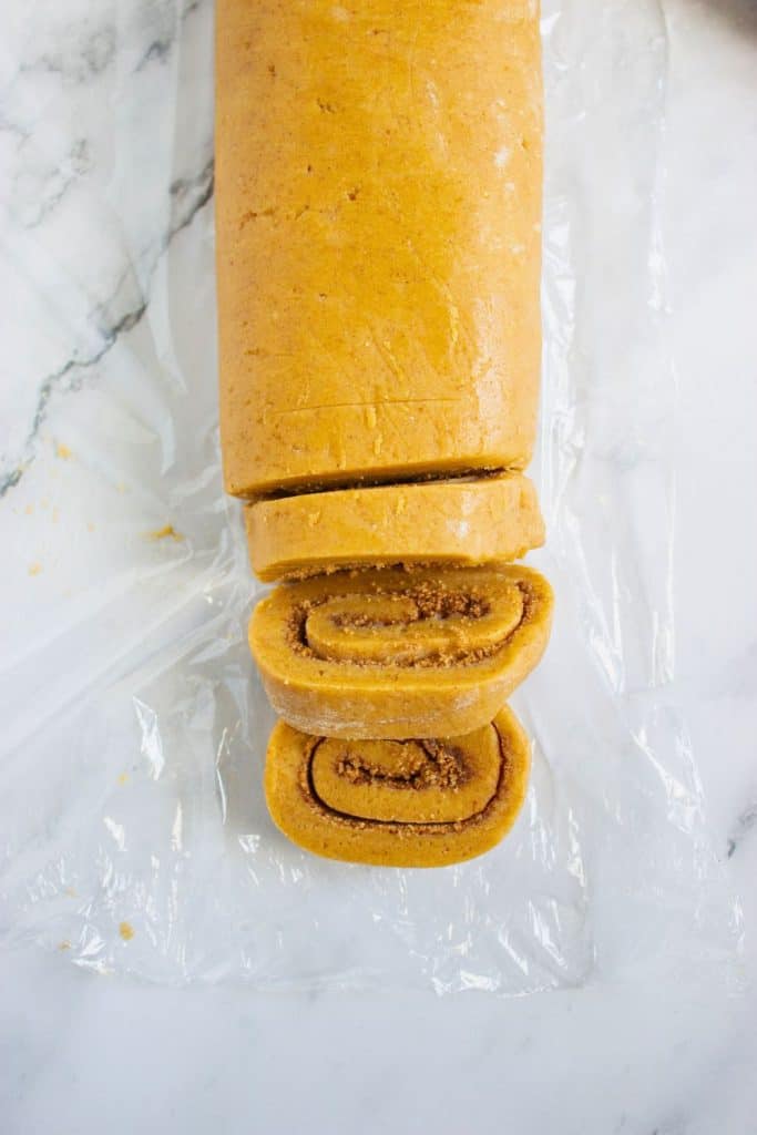 Rolled log of pumpkin cookie dough partially sliced into thick rounds, revealing a cinnamon sugar swirl inside each piece. The dough rests on plastic wrap over a marble surface, ready for baking.