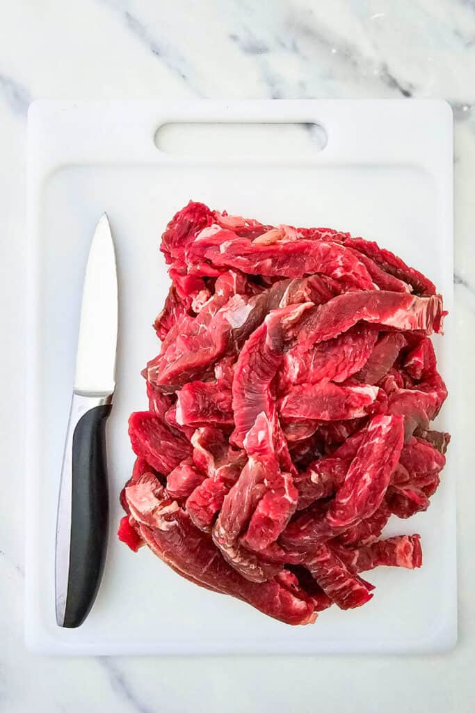 Overhead view of raw steak cut into thin strips, piled on a white cutting board. A kitchen knife rests beside the strips on a marble countertop.