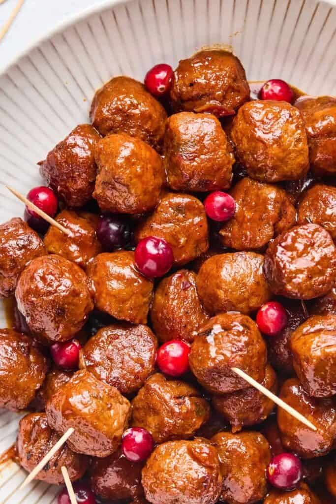 Slow cooker cranberry jalapeno meatballs coated in a glossy reddish brown sauce and served in a large white bowl. Fresh red cranberries are scattered among the meatballs with wooden toothpicks for serving.