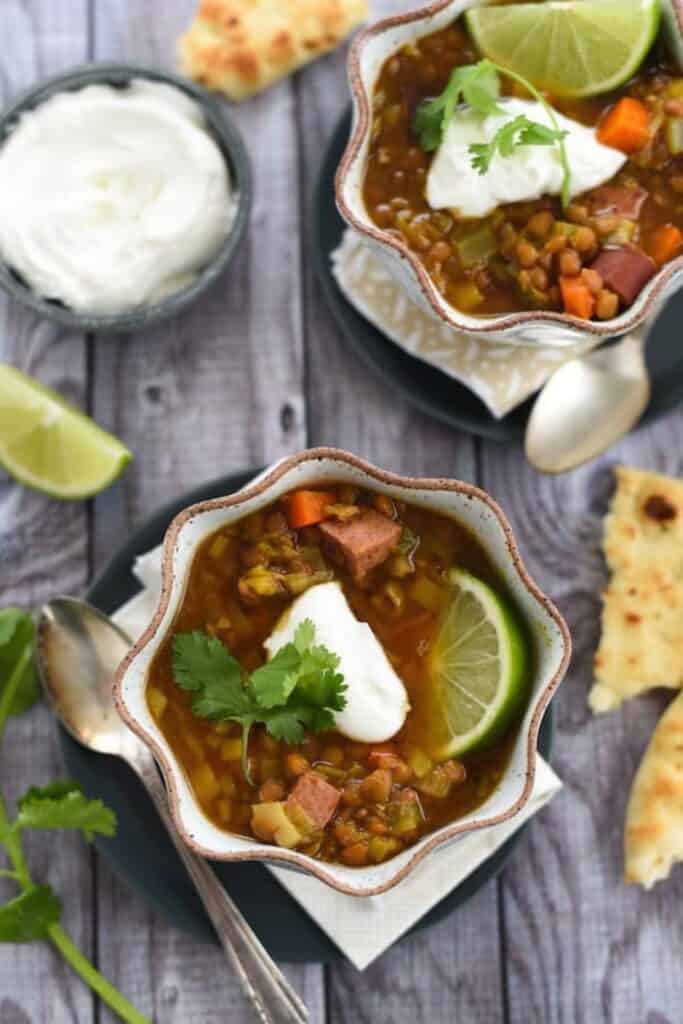 Close up of scalloped bowls with lential sausage soup topped with a dollop of sour cream and a lime slice. 