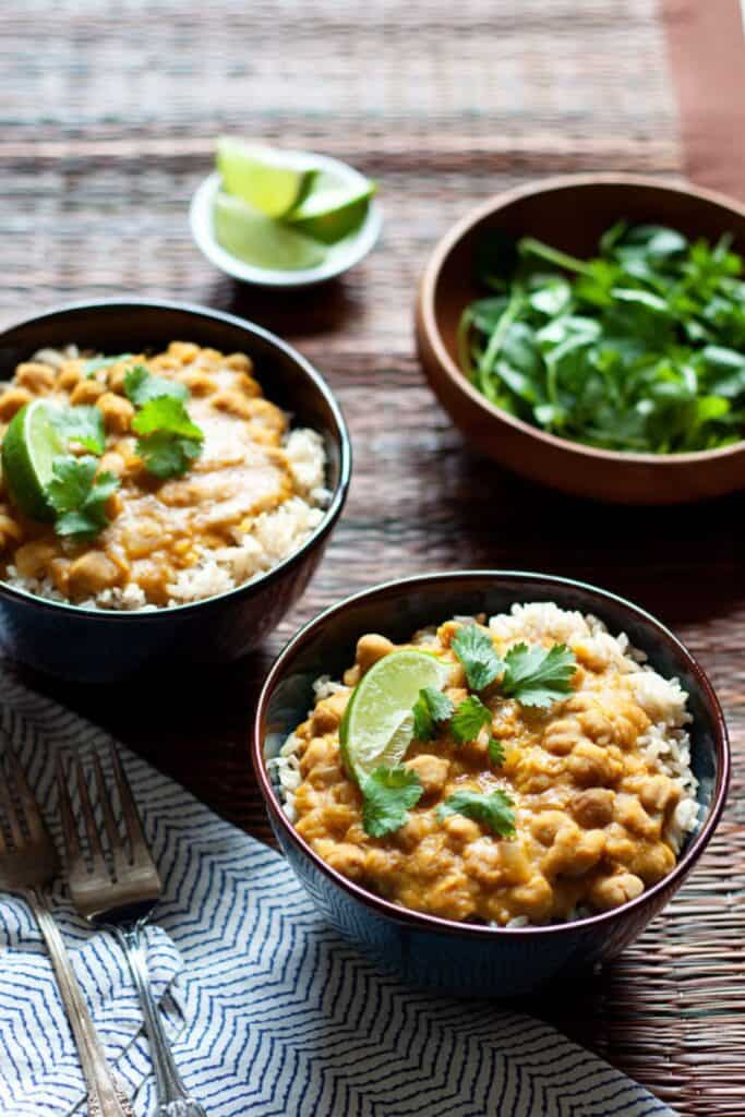 Two pottery bowls of pumpkin chickpea red lentil curry over rice, topped with lime slices and cilantro garnish.