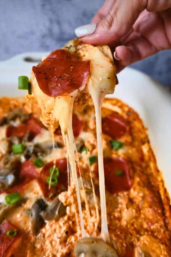 Cheesy easy game day dip being scooped with a chip, topped with pepperoni and green onions. Melted cheese stretches from the creamy baked dip in the dish below, making it a hearty and shareable football party snack.