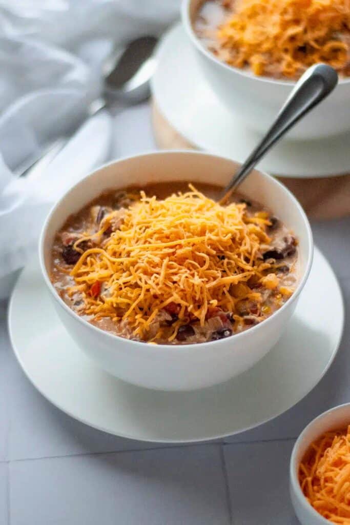 Bowl of Crockpot white chicken chili topped with a generous layer of shredded cheddar cheese melting into the creamy broth. The chili is filled with tender shredded chicken, beans, and diced vegetables, creating a thick and hearty texture.