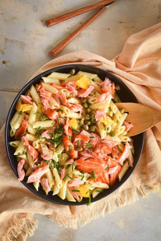 Bowl of smoked salmon pasta salad with penne pasta flaked smoked salmon capers orange segments and fresh chives tossed in a light dressing. Wooden chopsticks and a serving spoon sit beside the bowl on a soft cloth.