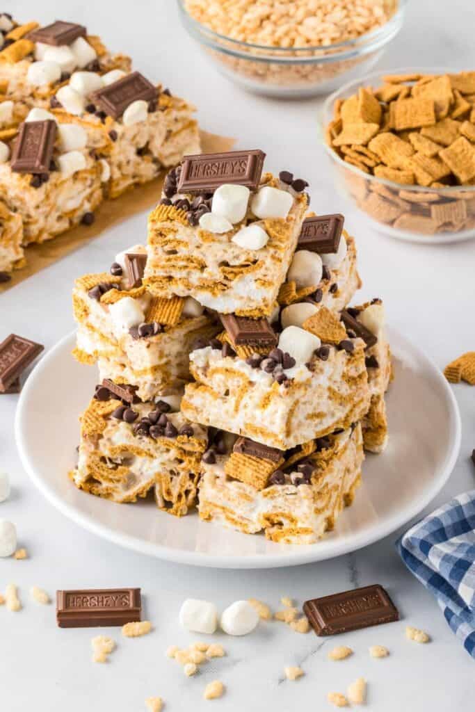 Stack of gooey S&rsquo;mores Rice Krispies treats topped with mini marshmallows, chocolate chips, graham cereal pieces, and chunks of chocolate bars. The no bake bars are layered with marshmallow cereal and finished with classic s&rsquo;mores toppings for an easy campout dessert.