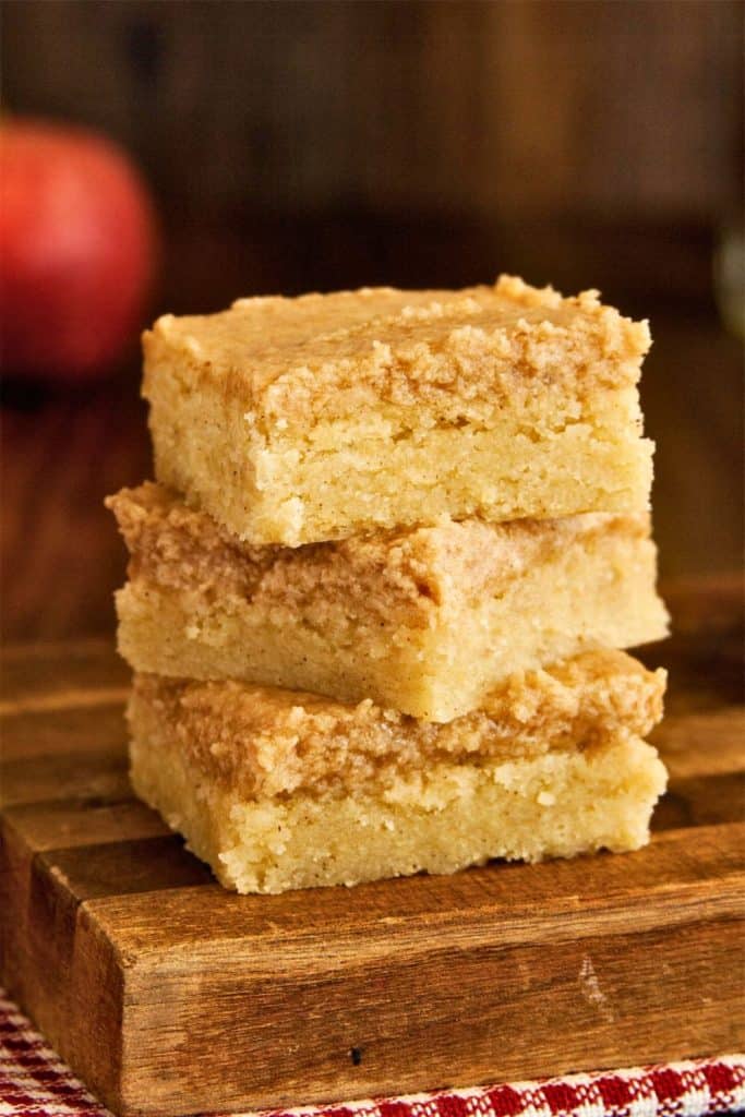 A stack of three snickerdoodle cookie bars with a golden, slightly crisp top and dense, moist interior, placed on a wooden board. The warm lighting and rustic background add a cozy, homemade feel to the fall dessert.
