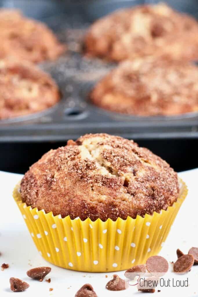 Close-up of a snickerdoodle muffin in a yellow polka dot wrapper, topped with a cinnamon-sugar crust and surrounded by scattered chocolate chips. The golden muffin is moist and fluffy, with other muffins visible in the background inside a baking tin.