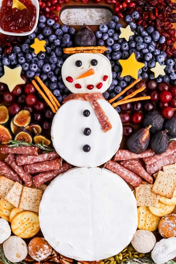 A Snowman charcuterie board featuring rounds of brie cheese stacked as the snowman’s body, decorated with blueberries, pomegranate seeds, a carrot nose, and pretzel stick arms, surrounded by festive fruits, meats, cheeses, crackers, and sweets.