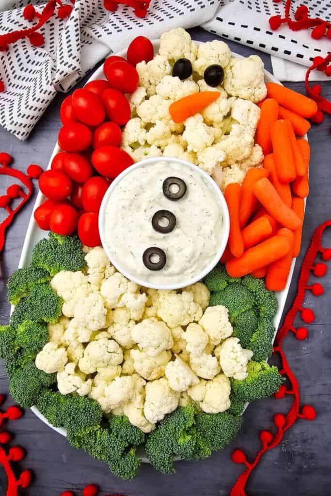 Snowman veggie platter made with cauliflower florets, cherry tomatoes, baby carrots, and broccoli arranged around a bowl of creamy dip. Black olives and a baby carrot create the snowman face and buttons for a festive Christmas appetizer.