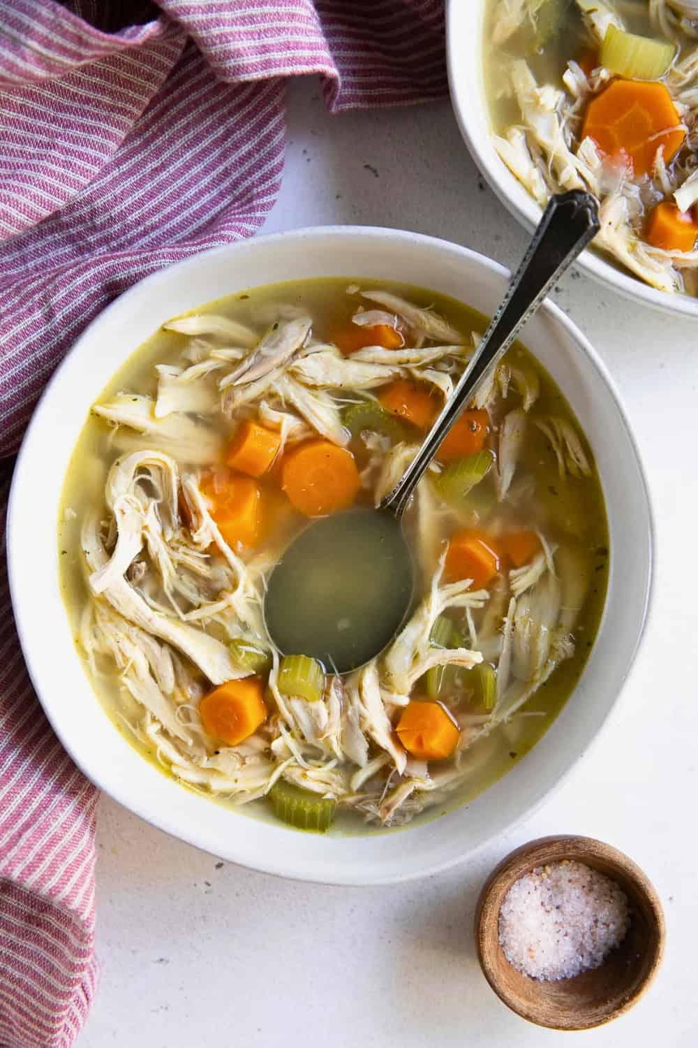 asy chicken soup with shredded chicken sliced carrots and celery is served in a light golden broth. A ladle rests in the bowl with a second serving of soup in the background.