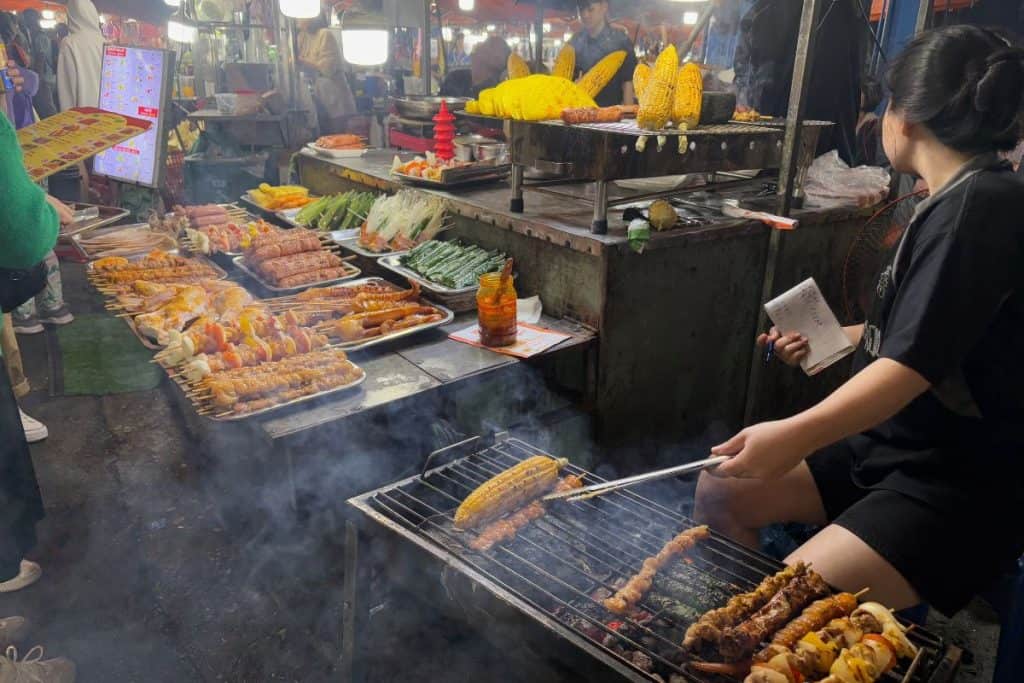A bustling food stall at Son Tra Night Market in Da Nang showcases trays of skewered meats, seafood, and vegetables ready for grilling. A vendor tends to the smoky charcoal grill, cooking corn and skewers while taking orders, as customers browse under the bright market lights in the lively evening atmosphere.