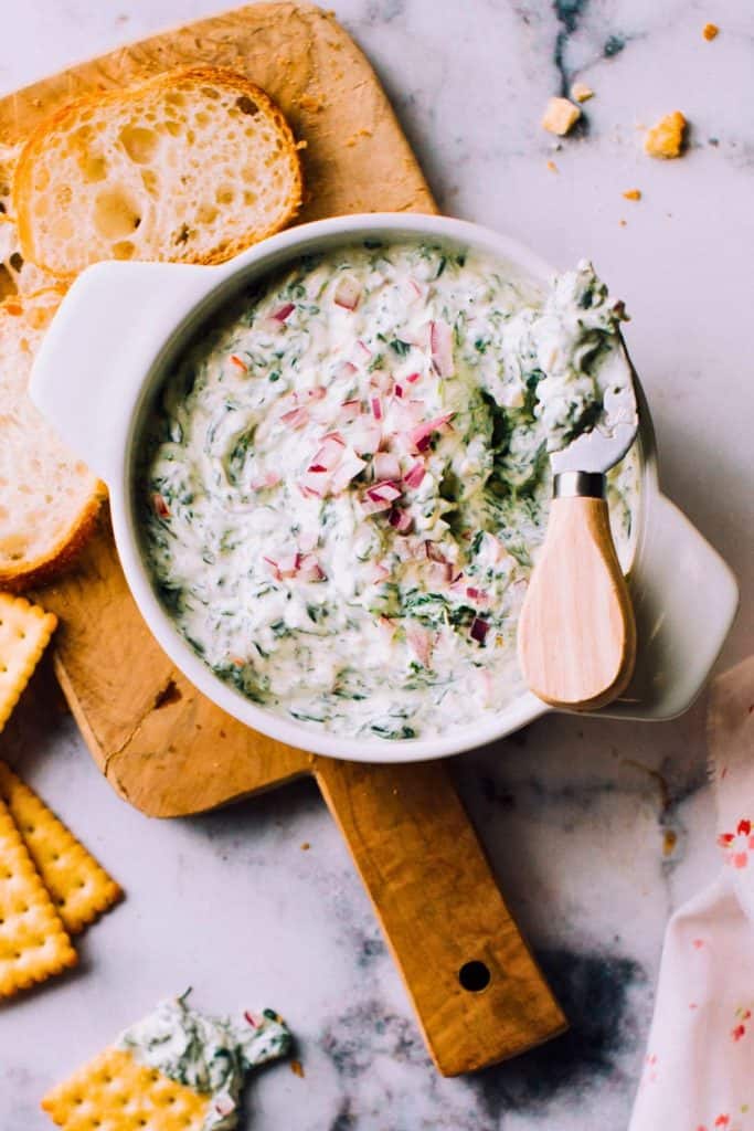 Creamy Sour Cream Spinach Dip in a white serving dish, garnished with diced red onions. Served with sliced bread and crackers on the side, for dipping and snacking.