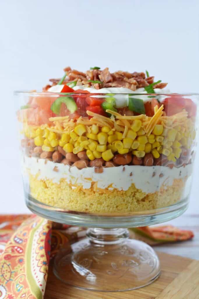 Layered Southern cornbread salad for a summer cookout is in a clear glass trifle dish, featuring distinct layers of crumbled cornbread, creamy dressing, pinto beans, corn, shredded cheddar cheese, chopped tomatoes, green bell peppers, and crispy bacon crumbles on top. The salad is set against a white background with a colorful paisley napkin nearby.
