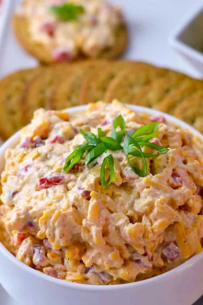 Southern Pimento Cheese Dip in a white bowl, topped with fresh sliced green onions. The creamy dip is filled with shredded cheddar, pimentos, and spices, served with crackers in the background.