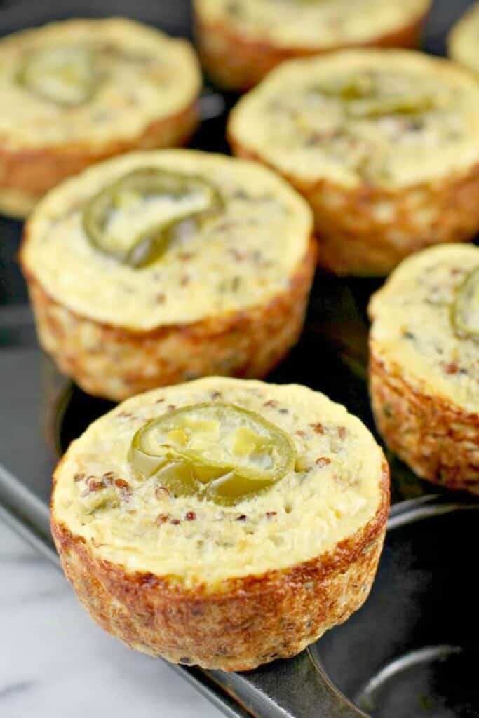 Southwest quinoa egg muffins with a golden baked exterior topped with jalapeno slices and visible quinoa throughout. These muffins sit in a muffin tin showing their firm texture and savory protein packed breakfast appeal.