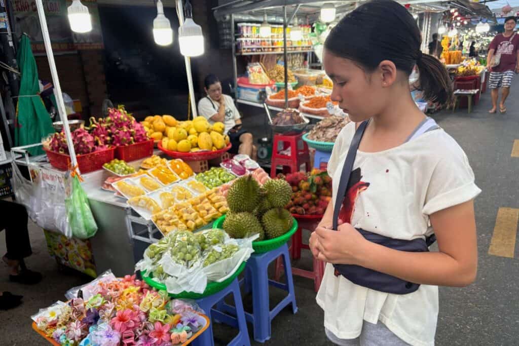  A young girl stands in front of a colorful fruit and souvenir stall at Phu Quoc Night Market, eyeing an array of tropical fruits like durian, dragon fruit, and rambutan, as well as packaged snacks and decorative flower hair clips. The bustling market scene is lit by bright hanging bulbs and filled with shoppers exploring the vibrant stalls.