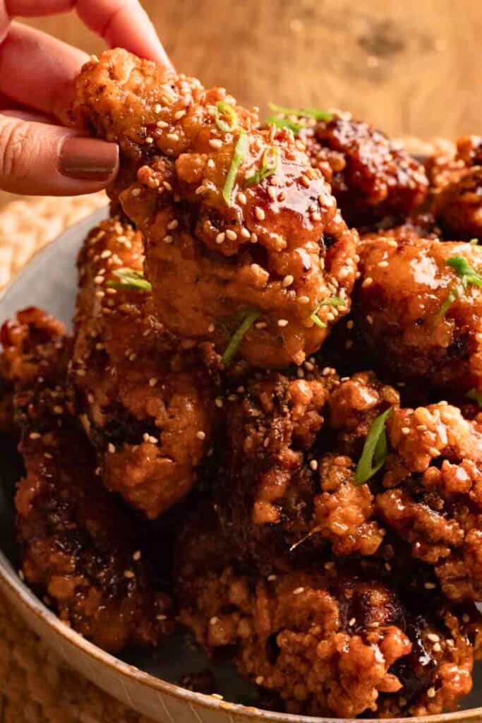 A hand picking up a crispy piece of soy garlic Korean fried chicken, coated in a glossy sauce and sprinkled with sesame seeds and green onion slices. The chicken is piled high in a bowl, with its golden-brown, crunchy texture.