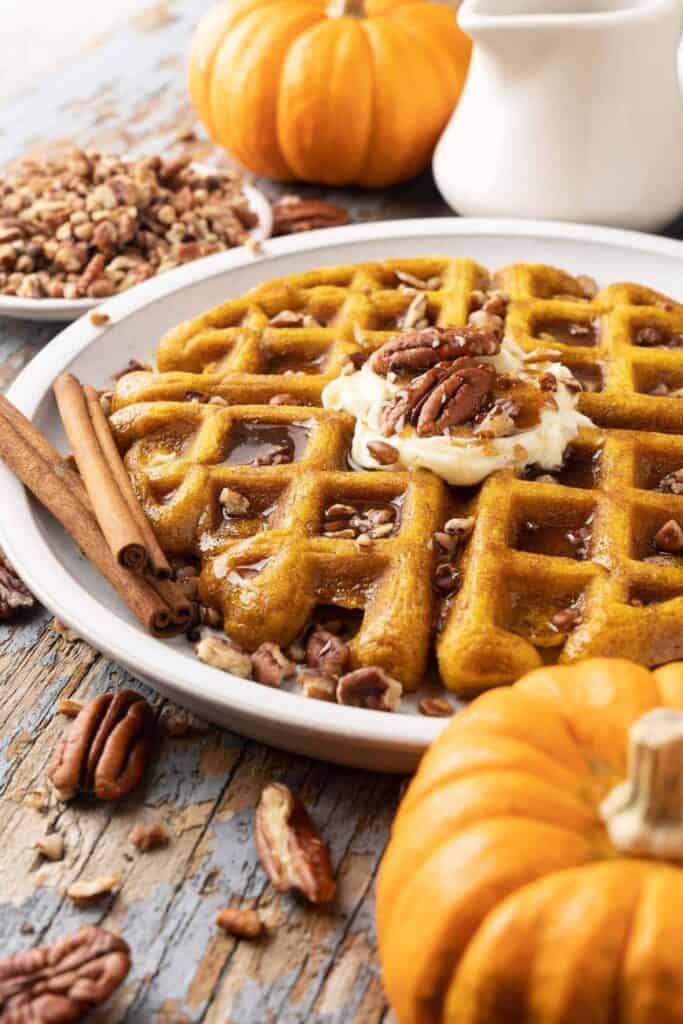 Pumpkin waffle on a plate topped with maple syrup, and pecans. 