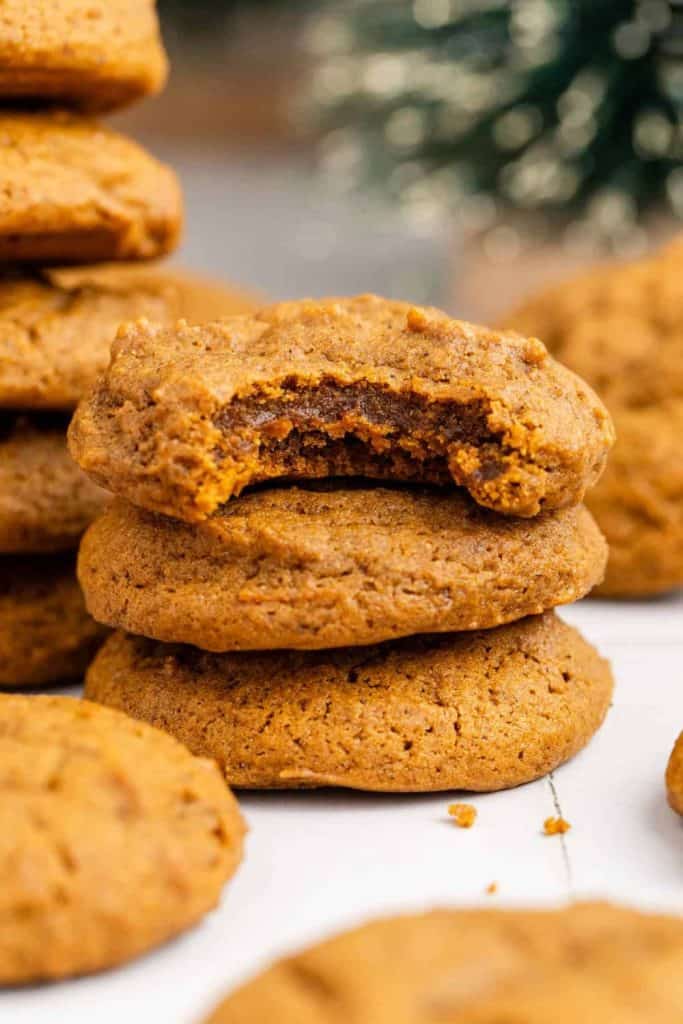 A stack of soft ginger molasses cookies with the top cookie showing a gooey, moist center from a bite taken out. The cookies are golden-brown with a cake-like texture, surrounded by more cookies and festive decor in the blurred background.