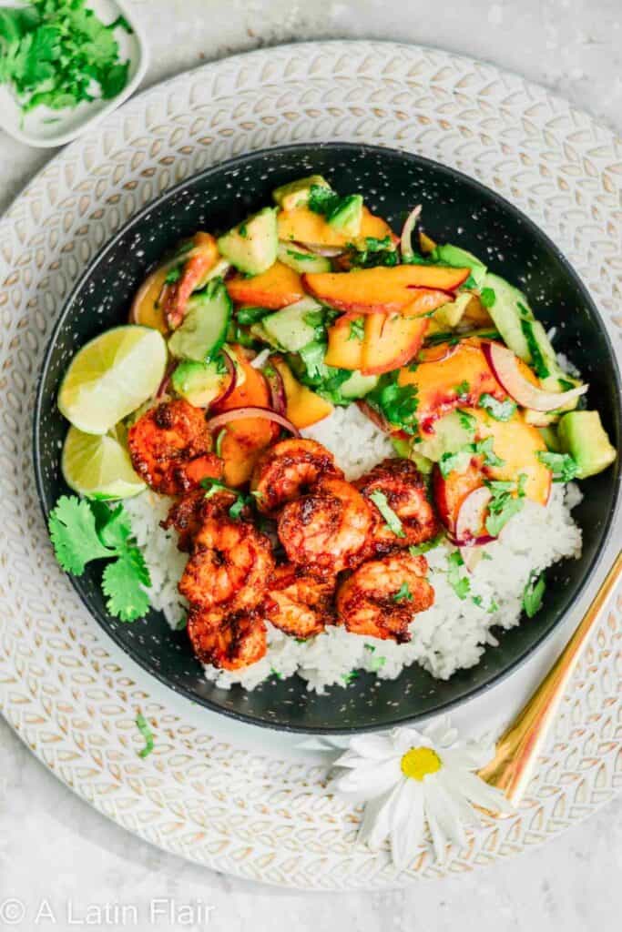 Spicy grilled shrimp served over white rice and topped with a fresh peach avocado salad made with sliced peaches, avocado, cucumber, red onion, and cilantro. Lime wedges on the side add a bright finishing touch to this colorful summer bowl that balances sweet fruit with smoky seafood.
