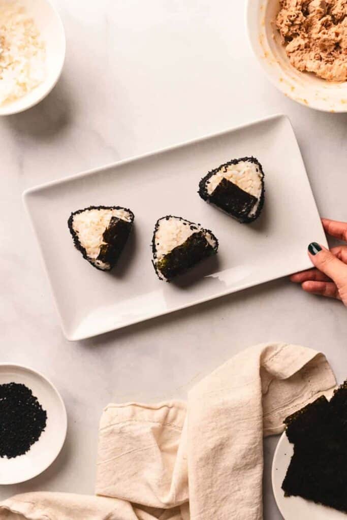 A hand holding a rectangular plate with three Japanese rice ball triangles "Onigiri" on it. 