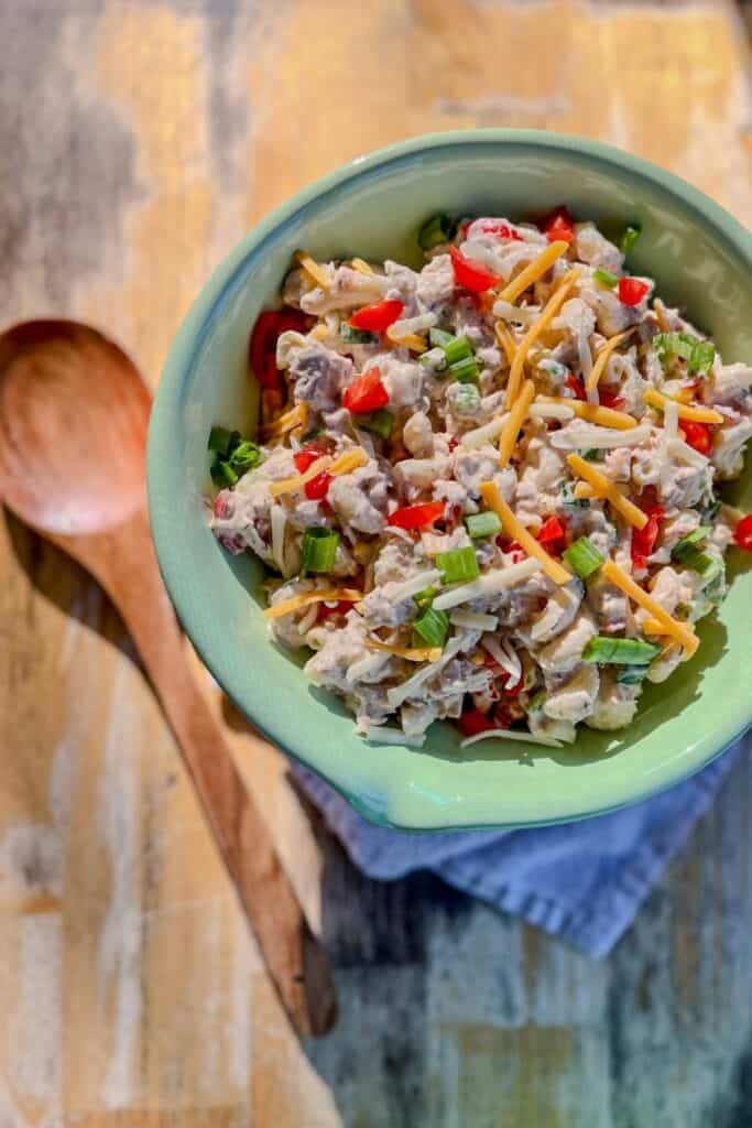 Bowl of spicy tuna pasta salad with small pasta tossed in a creamy dressing with chunks of tuna red peppers green onions and shredded cheese. The pasta salad is served in a green bowl on a wooden table with a spoon beside it.