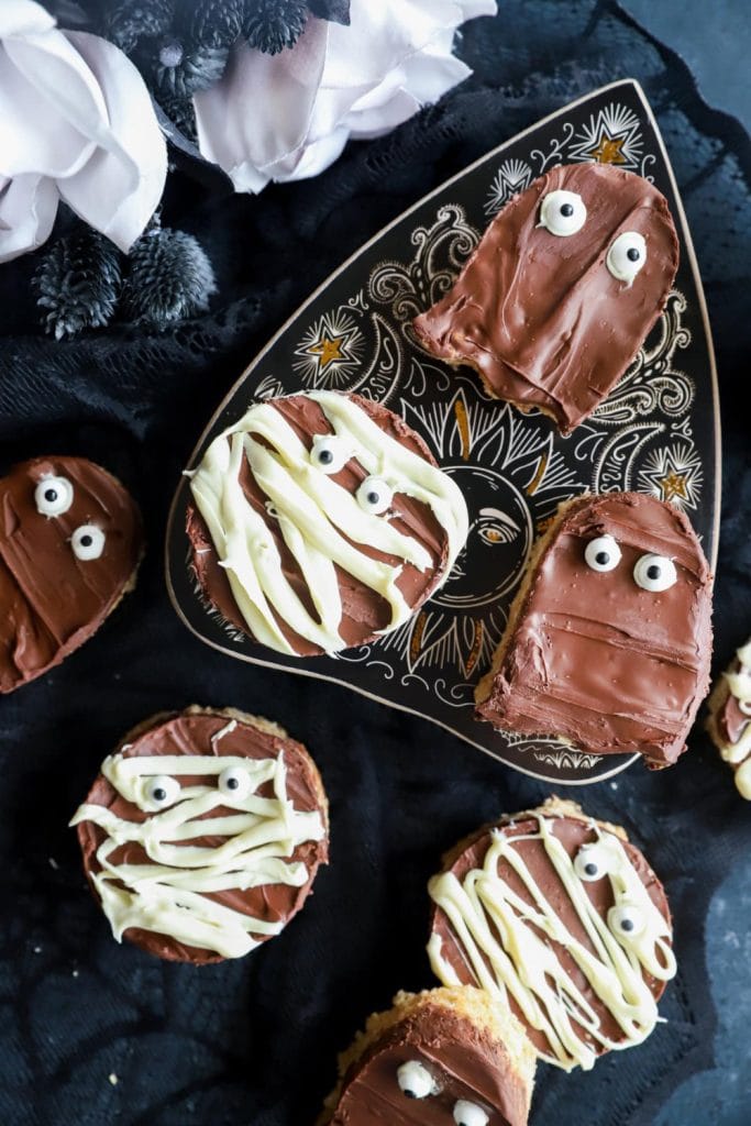 Spooky chocolate peanut butter no bake treats shaped like ghosts and mummies, topped with smooth milk chocolate, white chocolate drizzle bandages, and candy eyes, arranged on a decorative black and gold plate.