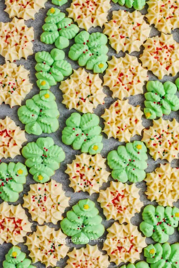  Overhead view of classic spritz Christmas cookies in festive shapes like green trees, wreaths, and buttery snowflakes are decorated with colorful holiday sprinkles, arranged in a pattern.  