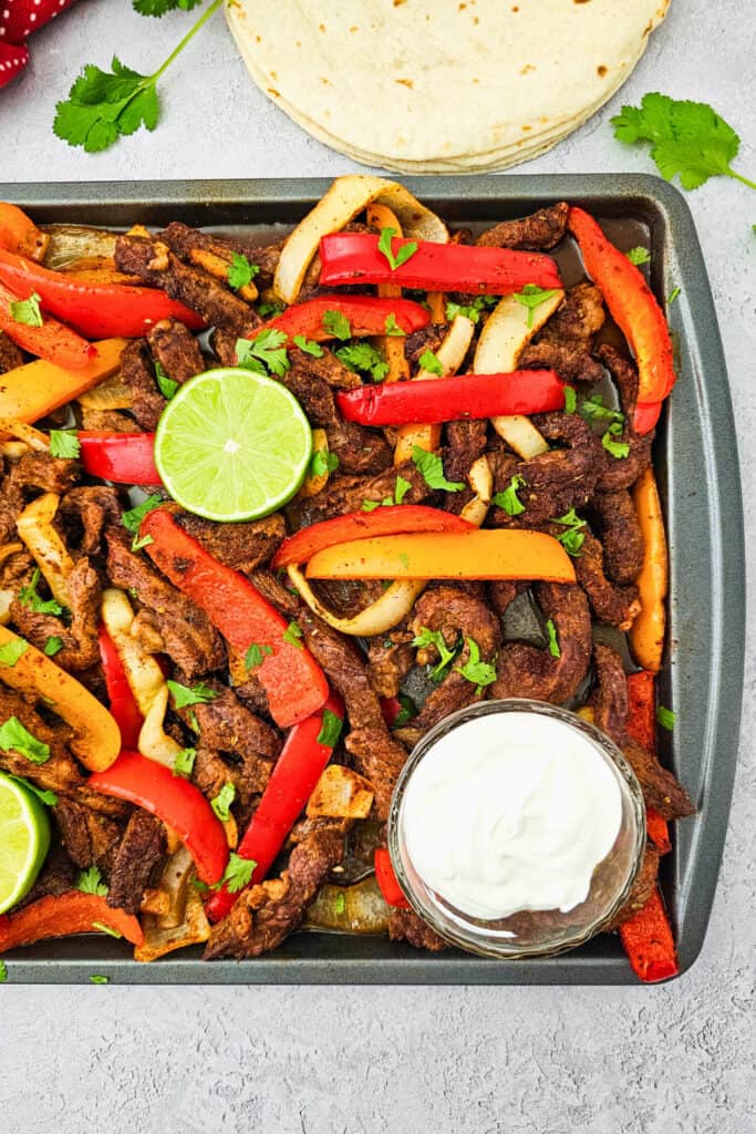 Overhead view of seasoned steak strips, roasted with red and orange bell peppers and onions for a colorful one pan dinner. The steak fajitas are topped with fresh cilantro and lime and served with a bowl of sour cream and flour tortillas.