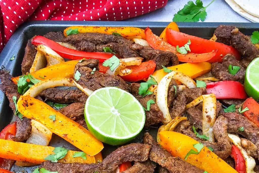 Seasoned strips of steak are roasted on a sheet pan with sliced red and orange bell peppers and onions. The steak sheet pan fajitas are garnished with fresh cilantro and bright green lime halves.