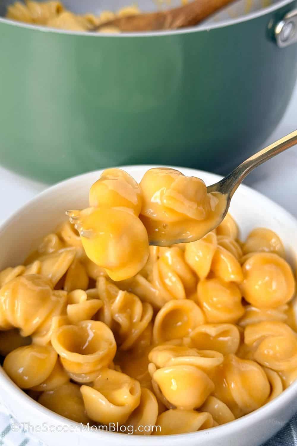 Spoon lifting creamy shell pasta coated in a smooth, rich cheese sauce above a bowl, with the cooking pot blurred in the background. This stovetop mac and cheese is extra cheesy, silky, and ready in one pot for a quick and easy comfort food dinner.