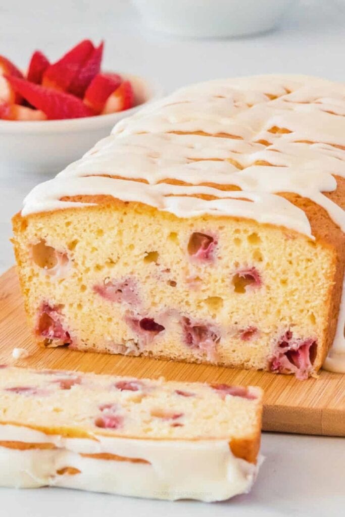 Loaf of strawberry bread topped with a thick white glaze and sliced to reveal juicy pieces of fresh strawberries baked into the soft crumb. The moist strawberry bread has a tender texture and sweet icing that drips slightly over the edges.