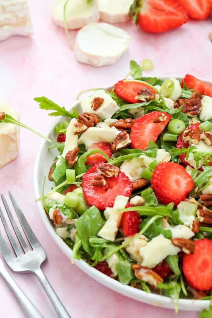 Fresh strawberry goat cheese salad served in a white bowl, topped with arugula, pecans, green onions, and creamy cheese chunks. Sliced strawberries and rounds of goat cheese are scattered in the background on a pink surface.