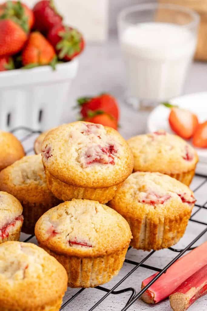 Golden strawberry rhubarb muffins cooling on a wire rack, with pieces of red strawberry and rhubarb baked into the tops. Fresh strawberries, rhubarb stalks, and a glass of milk surround the scene, adding a fresh, summery feel.