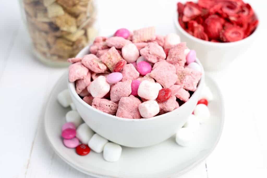 Small bowl of Valentine's Day Muddy Buddies with strawberry, white chocolate covered Chex cereal, M&Ms and mini marshmallows. With two small bowls of chex cereal and freeze dried strawberries in the background.